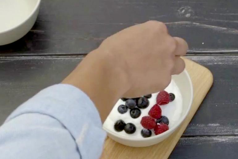A person's hand putting berries, blueberries and raspberries, into a bowl of yoghurt atop a wooden board, the person's hand obscures half the bowl.