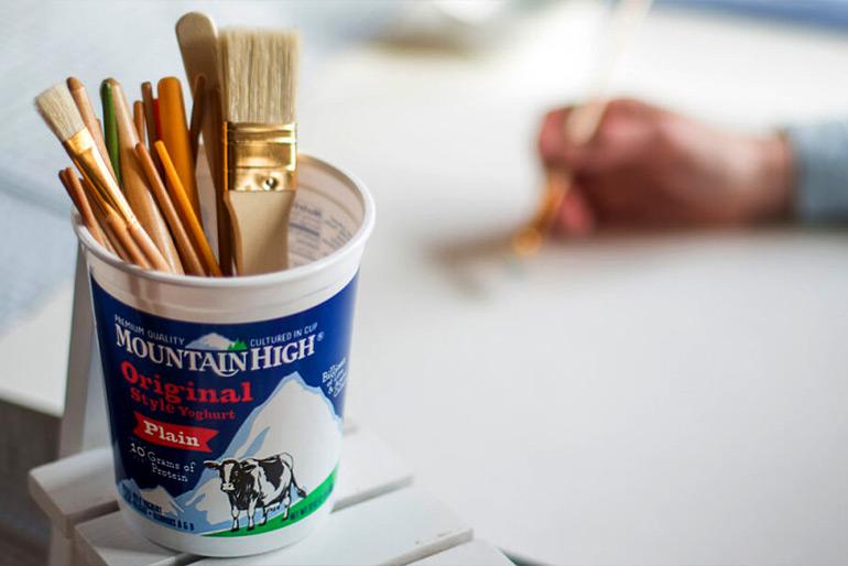A pot of Mountain High Plain Yoghurt that has been repurposed as a container for paintbrushes on various sizes, a hand is seen painting in the background.