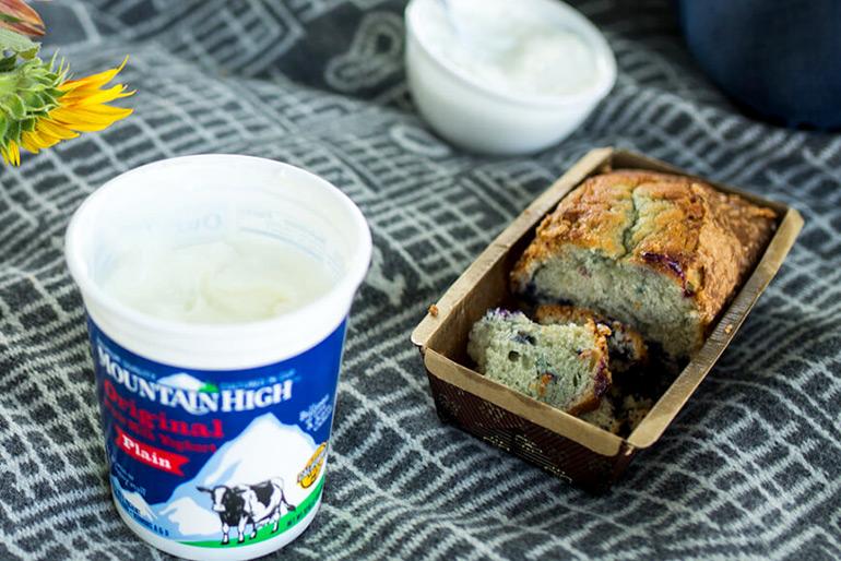 A pot of Mountain High Plain Yoghurt next to a tray with a half-eaten banana bread inside it. Behind the banana bread are a couple of bowls, and a flower.
