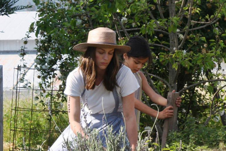 A mother and a daughter working on an allotment in a garden the mother is fiddling with plants, while the daughter drives a plank of wood into the ground.