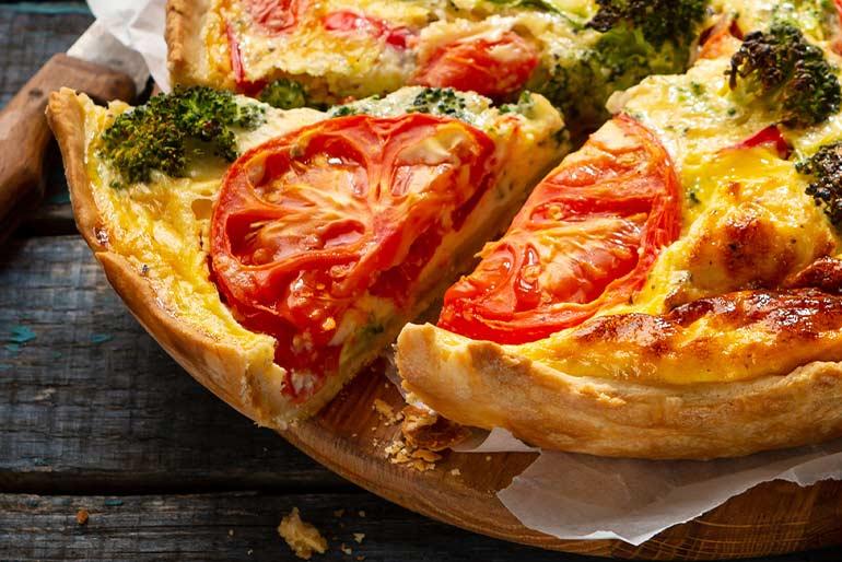 A quiche atop a wooden board made with salad tomatoes and broccoli, a slice of the quiche has been removed to show the cross section of the quiche.