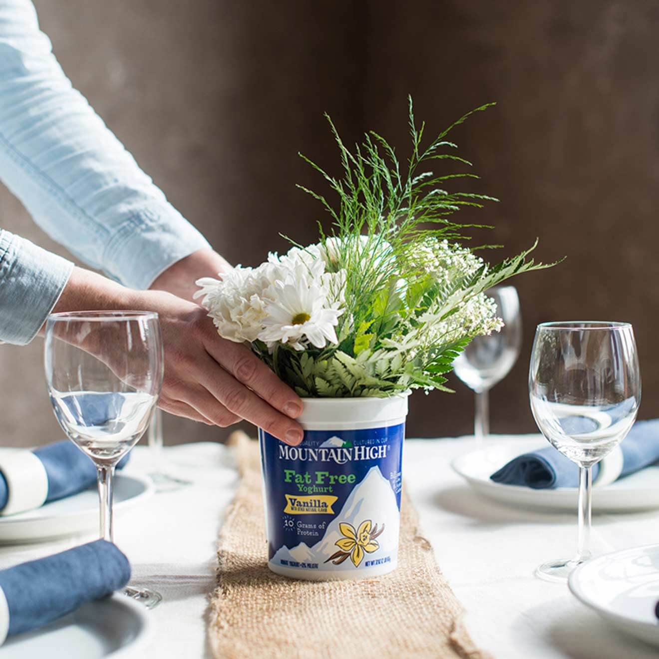 Hands placing a Mountain High Yoghurt container filled with white flowers on a table