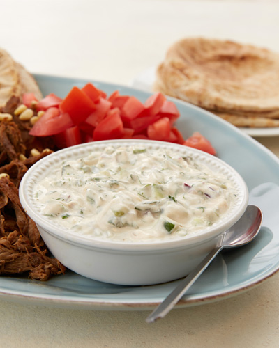 On a plate, there is sliced meat, tomatoes, and pita bread and a small bowl of Herbed Tahini Yoghurt Sauce, made with Mountain High Original Whole Milk Plain Yoghurt, tahini, lemon juice, cucumber, red onion, cilantro, mint, and garlic. Beside the bowl is a spoon. Next to the plate is a smaller plate of pita bread.